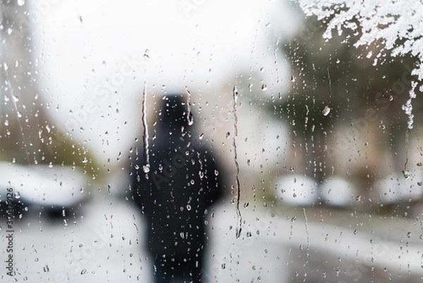 Obraz Unfocused silhouette man in a winter jacket with a hood behind car window with melted snow texture. Winter background