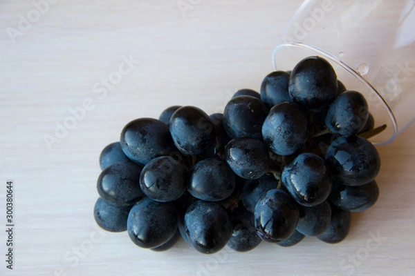 Obraz Bunches of fresh ripe red grapes and glass for wine on a white wooden background, top view, copy space. Table setting. Beautiful background with a branch of blue grapes. Red wine grapes.