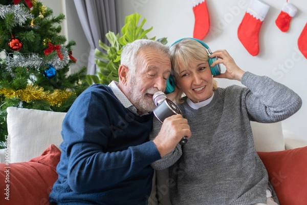Fototapeta senior elderly old man and woman happy to sing song and listening with headphone together in living room that decorated with christmas tree, retirement lovely lifestyle concept