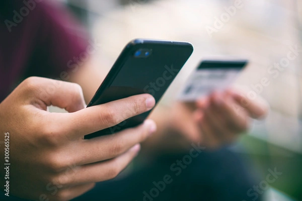 Fototapeta Close-up human hands with mobile phone and credit card. Person holds smartphone and bank card, closeup shot. On foreground male hands hold cell phone and card. Soft focus effect. Outdoors. In the city