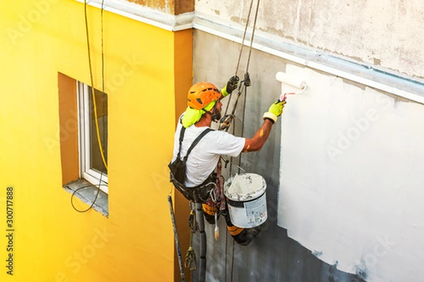 Fototapeta Industrial rope access worker hanging from the building while painting the exterior facade wall. Industrial alpinism concept image. Top view