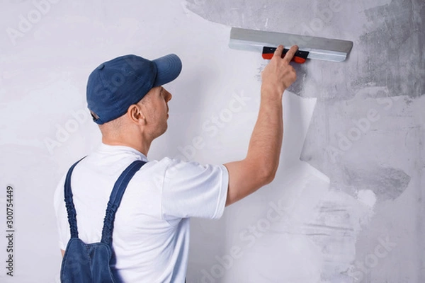 Fototapeta Worker in blue overalls and cap plastering a wall with finishing putty using a putty knife. Repair work and construction concept. Close-up