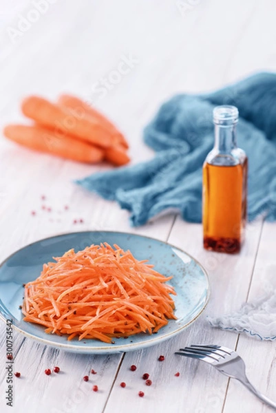 Fototapeta Grated carrot salad on blue plate, peeled carrots, bottle with olive oil, pink peppercorns, blue towel and a fork on the white wooden table. Organic food concept. High key vertical image, copy space
