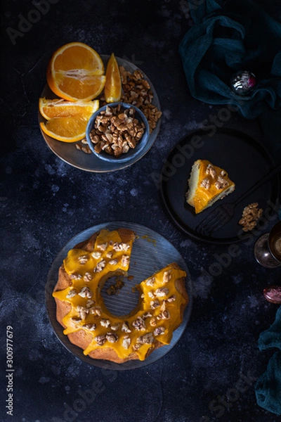 Fototapeta Bundt cake, orange cake with walnuts and yellow sugar icing with turmeric spice. Dark background. View from above. With a cut piece