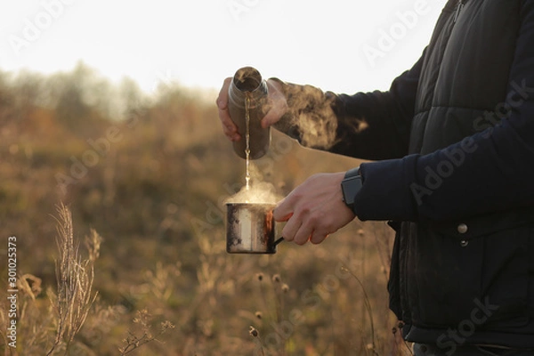 Obraz A woman pours a hot drink from a thermos into a mug. Refreshment while hiking. Clink glasses of tea in the morning at dawn, three people drink tea. Metal cups on the nature. Hands warm hot coffee