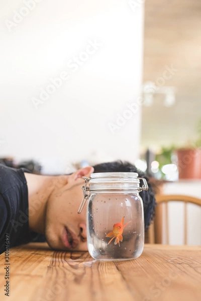Fototapeta A young boy lying down on the wooden table look at the gold fish in the jar.