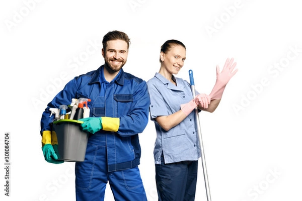 Obraz Attractive young woman and man  in cleaning uniform and rubber gloves holding  a mop and  a bucket of cleaning products in his hands, isolated on white background.