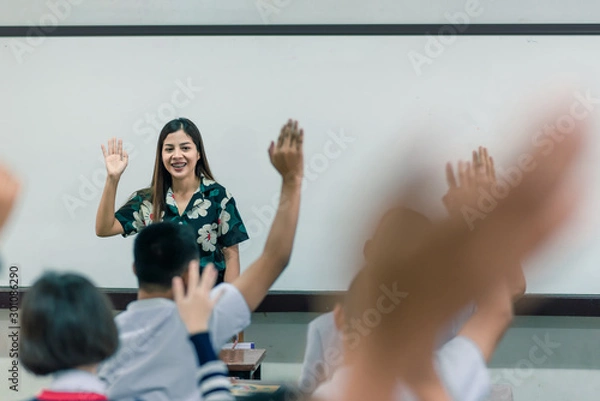 Fototapeta An smiling Asian female high school teacher teaches the white uniform students in the classroom by asking questions and then the students raise their hands for answers.