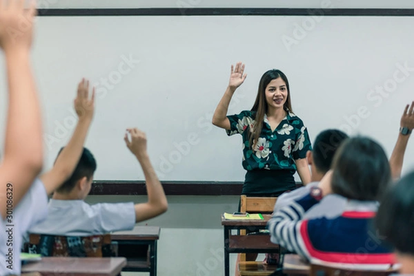 Fototapeta An smiling Asian female high school teacher teaches the white uniform students in the classroom by asking questions and then the students raise their hands for answers.