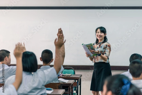Fototapeta An smiling Asian female high school teacher teaches the white uniform students in the classroom by asking questions and then the students raise their hands for answers.