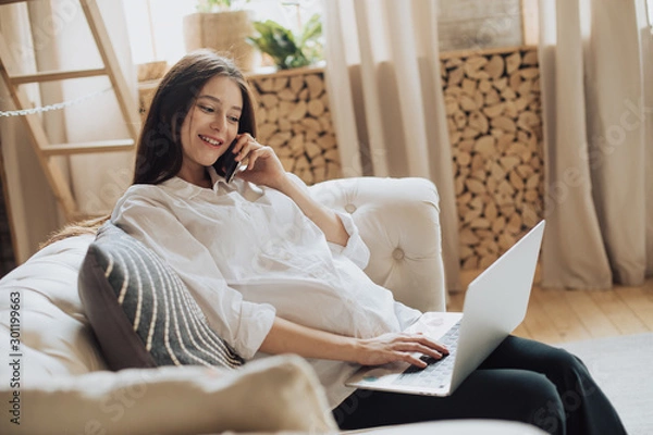 Fototapeta Young cheerful pregnant woman sit on couch and talk on phone. Also working remote using laptop. Smile and look at screen. Sit alone in room. Build her own business. Expect a baby in future.