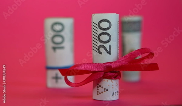 Fototapeta Rolled banknotes of 200 Polish zloty tied with red ribbon concept of economical / financial gift, behind two rolls of 100 zloty in soft focus, isolated on pink background close up