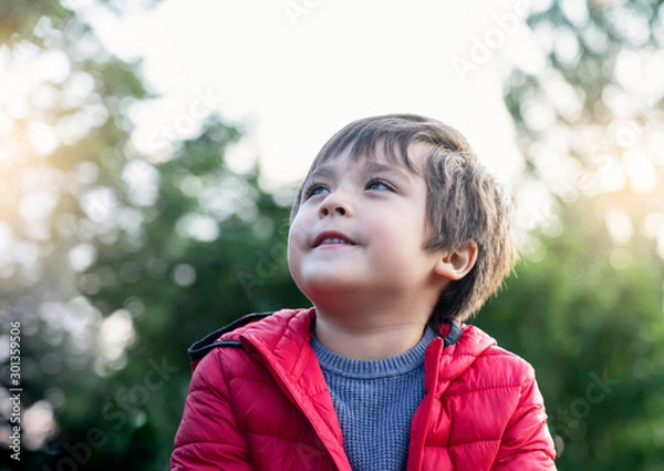Fototapeta Candid shot of happy little kid looking up with smiling face sitting in the park,Cropped shot of child boy looking up to sky relaxing on his own with blurry natural green forest backgroud