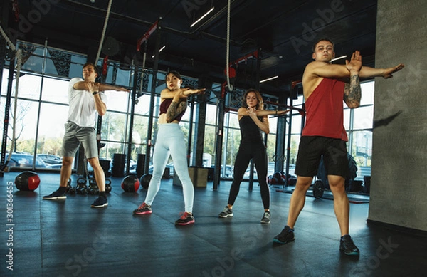 Fototapeta A group of muscular athletes doing workout at the gym. Gymnastics, training, fitness workout flexibility. Active and healthy lifestyle, youth, bodybuilding. Training in lunges and stretching exercises