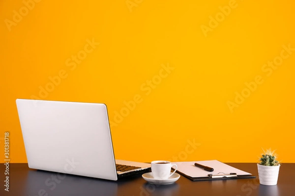 Fototapeta Back of the laptop computer, various supplies on wooden desk over bright yellow wall background with a lot of copy space for text. Creative workspace. Close up shot of table w/ computer & stationery.