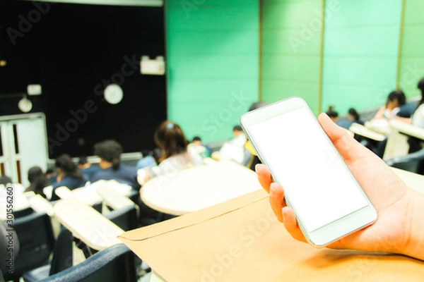 Fototapeta Female using white mobile phone with a blank screen in the classroom and blur background of students during the study, quiz, test or exams from a teacher in a large lecture room/University classroom.