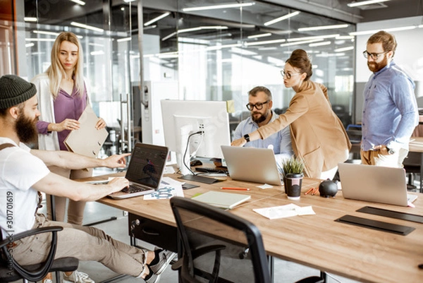 Fototapeta Grupa różnorodnych kolegów pracujących na komputerach w nowoczesnym biurze lub coworkingu, brodaty hipster jako programista piszący kod
