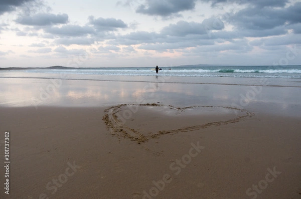 Obraz Heart drawn in the sand with people surfing in the background