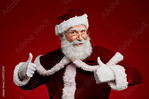Obraz Waist up portrait of cheerful Santa Claus showing thumbs up while standing against red background in studio, copy space