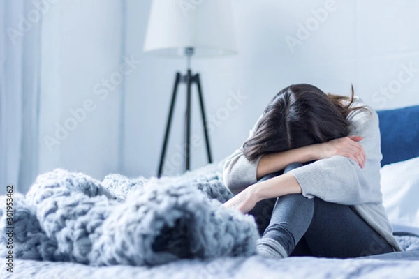 Fototapeta Młoda kobieta w szarym ubraniu siedzi skulona na łóżku w domu. Zdenerwowana brunetka dziewczyna cierpi, płacze. Depresja z powodu samotności, stresu, problemów. Koncepcja zaburzeń psychicznych.