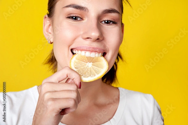 Obraz Girl bites a slice of lemon. Close-up, on a bright yellow background. Healthy nutrition, cold prevention, vitamins.
