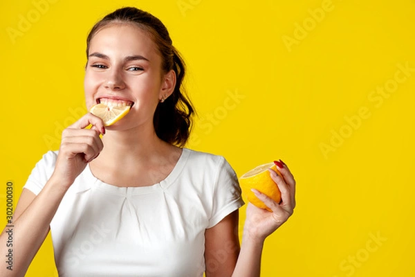 Fototapeta A girl in a white T-shirt is eating a lemon, on a bright yellow background. Free space for text.