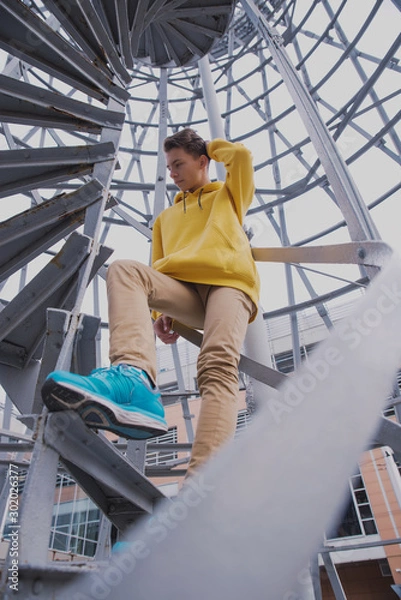 Obraz A young guy in a yellow hoodie and turquoise sneakers stands on a spiral staircase. Teen jumps off with a beautiful spiral staircase in the beautiful architecture
