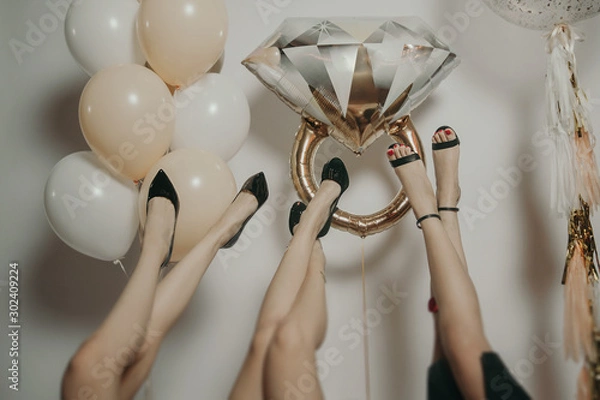 Fototapeta nogi dziewcząt w czarnych butach leżą do góry nogami na jasnym tle ze złotymi i białymi balonami i pierścieniem balonu dla panny młodej