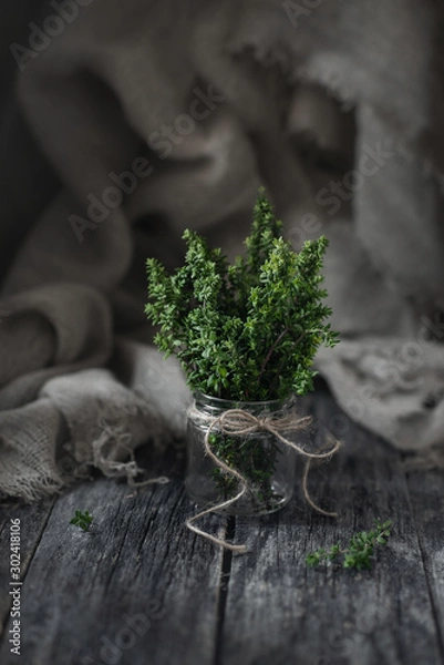 Fototapeta A thyme bundle in a glass jar and scattered thyme sprigs on wooden table on sackcloth background. Magic light, vertical orientation, copy space