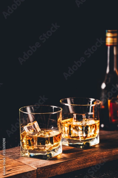 Fototapeta Two glasses of whiskey with ice cubes served on wooden board. Dark background, glossy golden color hard liquor on countertop with highlight. Night life bar relaxation concept