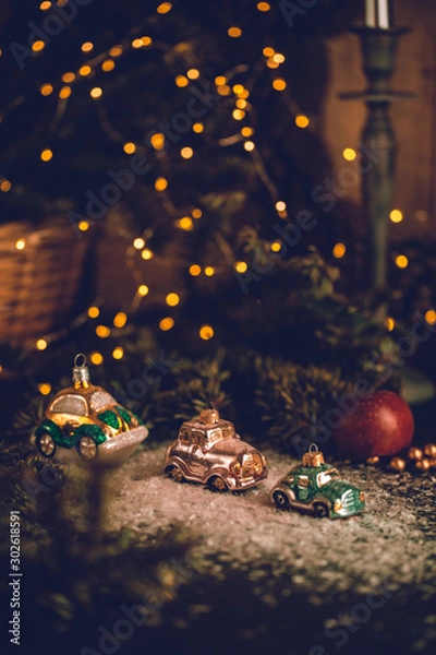 Fototapeta Christmas tree ornaments, tiny car on snowy table under the fir tree and bokeh lights with white candle in the background. Beautiful mysterious village decoration, greeting card, new year celebration 