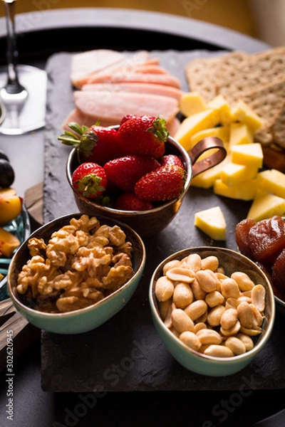 Fototapeta Cheese and fruits plate with glass of white wine on round tray, colorful and delicious charcuterie with snacks and wineglass for romantic evening, wine tasting, selective focus.