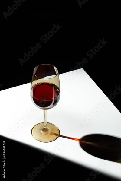 Fototapeta Object shot of a golden wineglass, staying on a white table. The light falls on the glass, the glass casts a shadow. The glass is half full of wine.