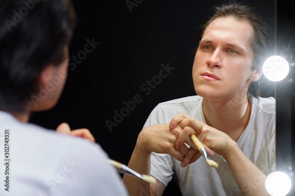 Obraz the actor sits in front of a mirror in a white t-shirt in a dark room and looks at his reflection, holding a brush for applying makeup to his face