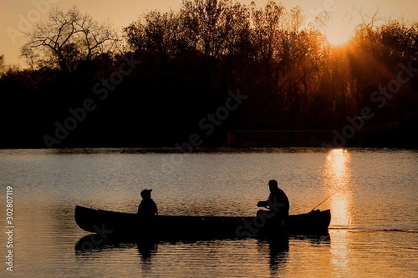 Fototapeta Close-Up Silhouette of a Man and a Child Together in a Canoe on a Lake in the Evening - with a Vibrant Orange Sunset in the Background
