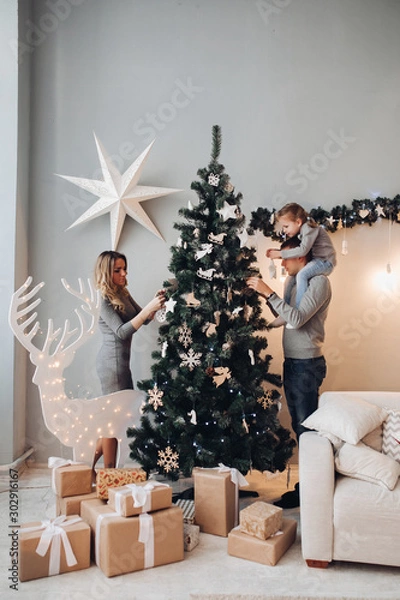 Obraz Full length stock photo of mother, father and daughter on father s shoulders decorating beautiful green Christmas tree with toys and decorations. Boxes of presents under the tree.