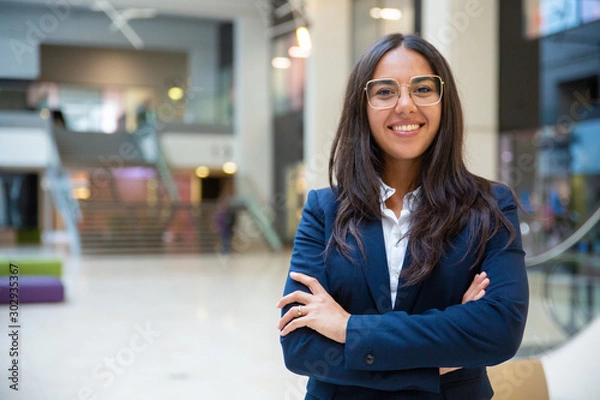 Fototapeta Happy successful businesswoman posing in office hall. Young Latin woman wearing formal suit and glasses, standing for camera with arms folded, smiling. Business portrait concept