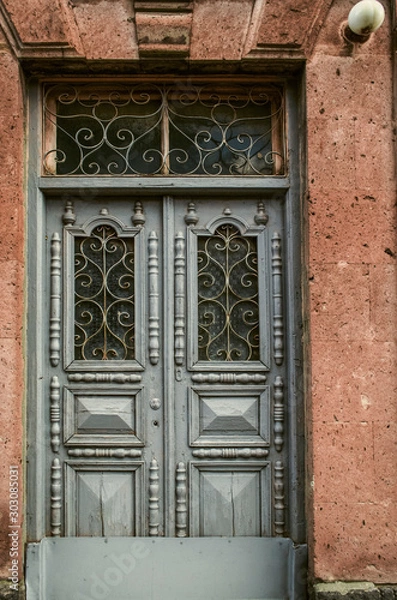 Fototapeta A light blue double wooden door with carved details is covered with openwork wrought iron bars on the windows of the door at the facade of the stone building