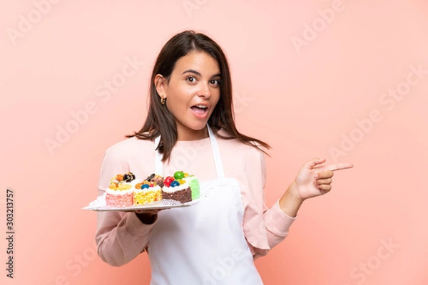 Fototapeta Young girl holding lots of different mini cakes over isolated background surprised and pointing finger to the side