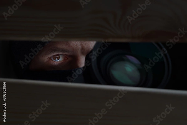 Fototapeta Male eye and camera close-up. Recording to a hidden camera. Private detective with camera looks at a cleft. Espionage background. The concept of eavesdropping, espionage, gossip and the yellow press