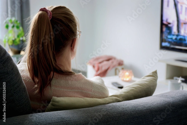 Fototapeta A young woman from behind with ponytail and glasses watching TV while sitting on sofa in living room at home. Woman binge watching tv via online streaming platform