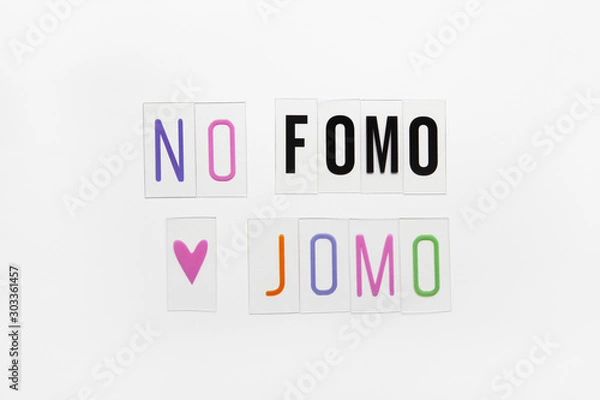Fototapeta Abbreviation words FOMO, JOMO on transparent plastic on white background. FOMO means Fear Of Missing Out. JOMO - Joy Of Missing Out. Opposition, choice, social problem, digital detox. Flat lay