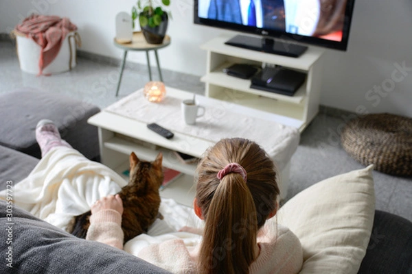 Obraz Domestic life with pet. A young woman is sitting on the couch with her cat on her lap in the living room. She watches TV while stroking her cat. Woman binge watching tv via online streaming platform.