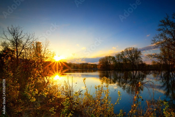 Fototapeta Long exposure sunset or sunrise over river side of Mississippi and the sun is reflex with water surface and distribution sun rays and small trees in foreground and clear blue sky