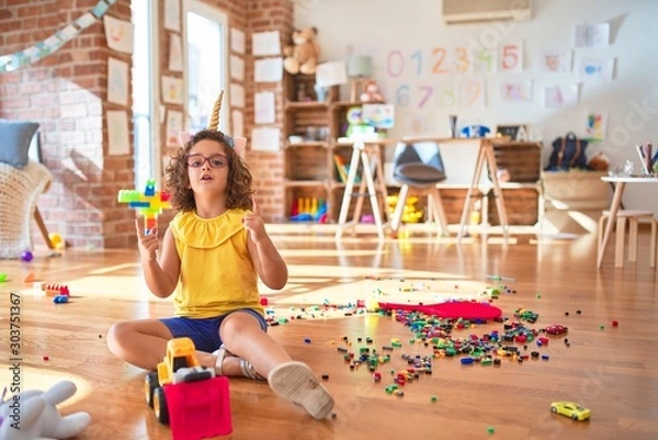 Fototapeta Beautiful toddler wearing glasses and unicorn diadem sitting on the floor at kindergarten amazed and surprised looking up and pointing with fingers and raised arms.