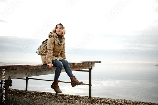 Fototapeta young girl in a warm jacket sits on a pier with legs dangling and look to the side