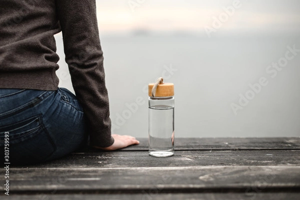 Fototapeta glass reusable bottle for water, stands on a wooden pier against the background of a cloudy sky next to a sitting figure of a girl