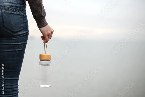 Fototapeta glass reusable water bottle hanging in a female hand on a background of cloudy sky
