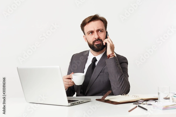 Fototapeta Severe young bearded businessman with short haircut and lush beard sitting at working table, having phone conversation while drinking coffee, isolated over white background