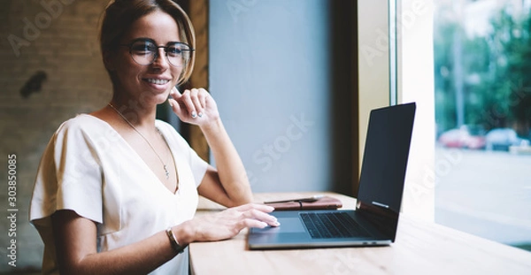 Fototapeta Portrait of cheerful millennial woman in trendy spectacles looking at camera with cute smile on face during blogging via laptop computer, positive hipster girl enjoying distance job in cafeteria